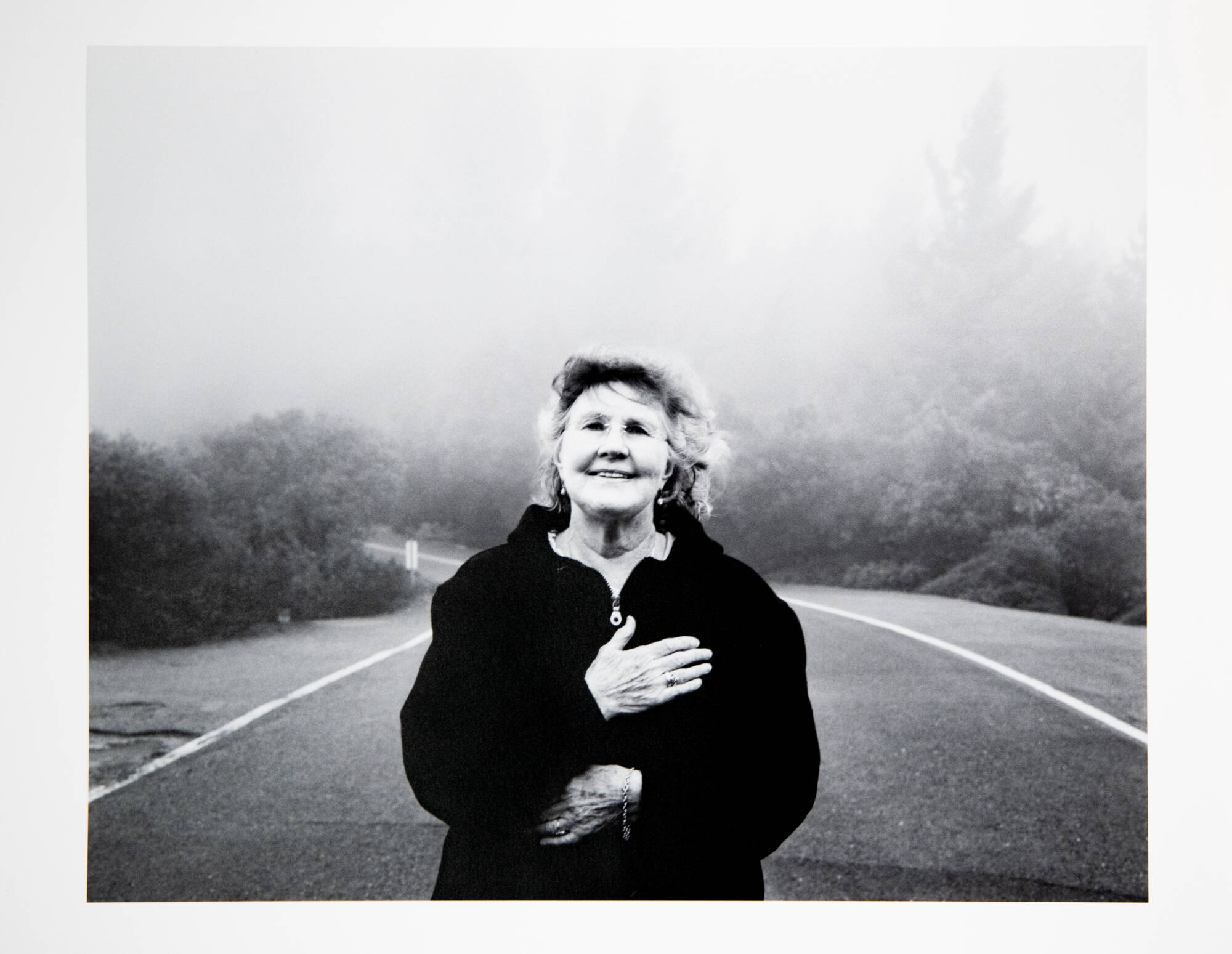 A black and white photograph of a woman with her hand over her heart. She is standing on a road with trees behind her.